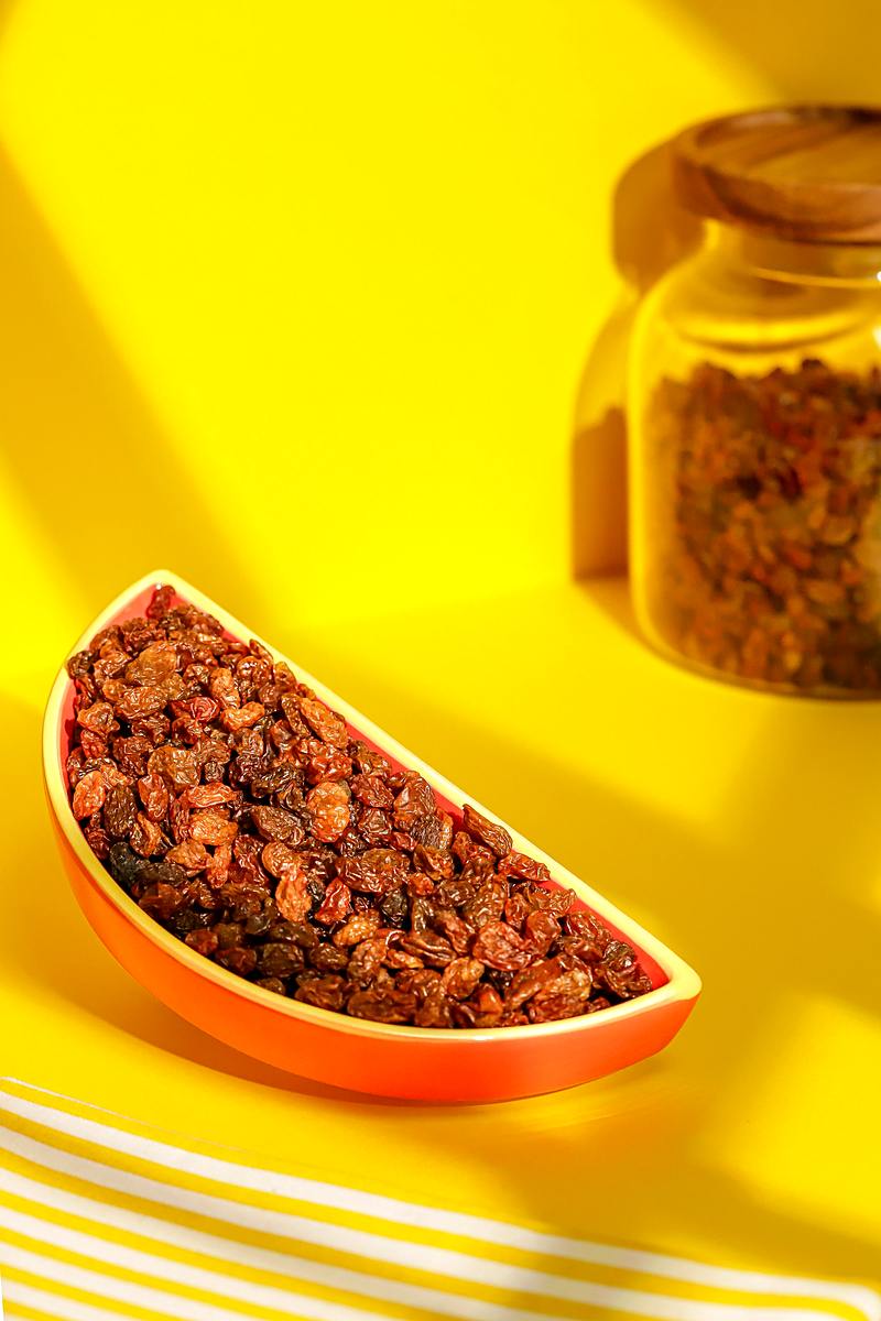 Raisins secs sultanine dans une coupelle allongée orange, sur fond jaune, avec un bocal en arrière-plan